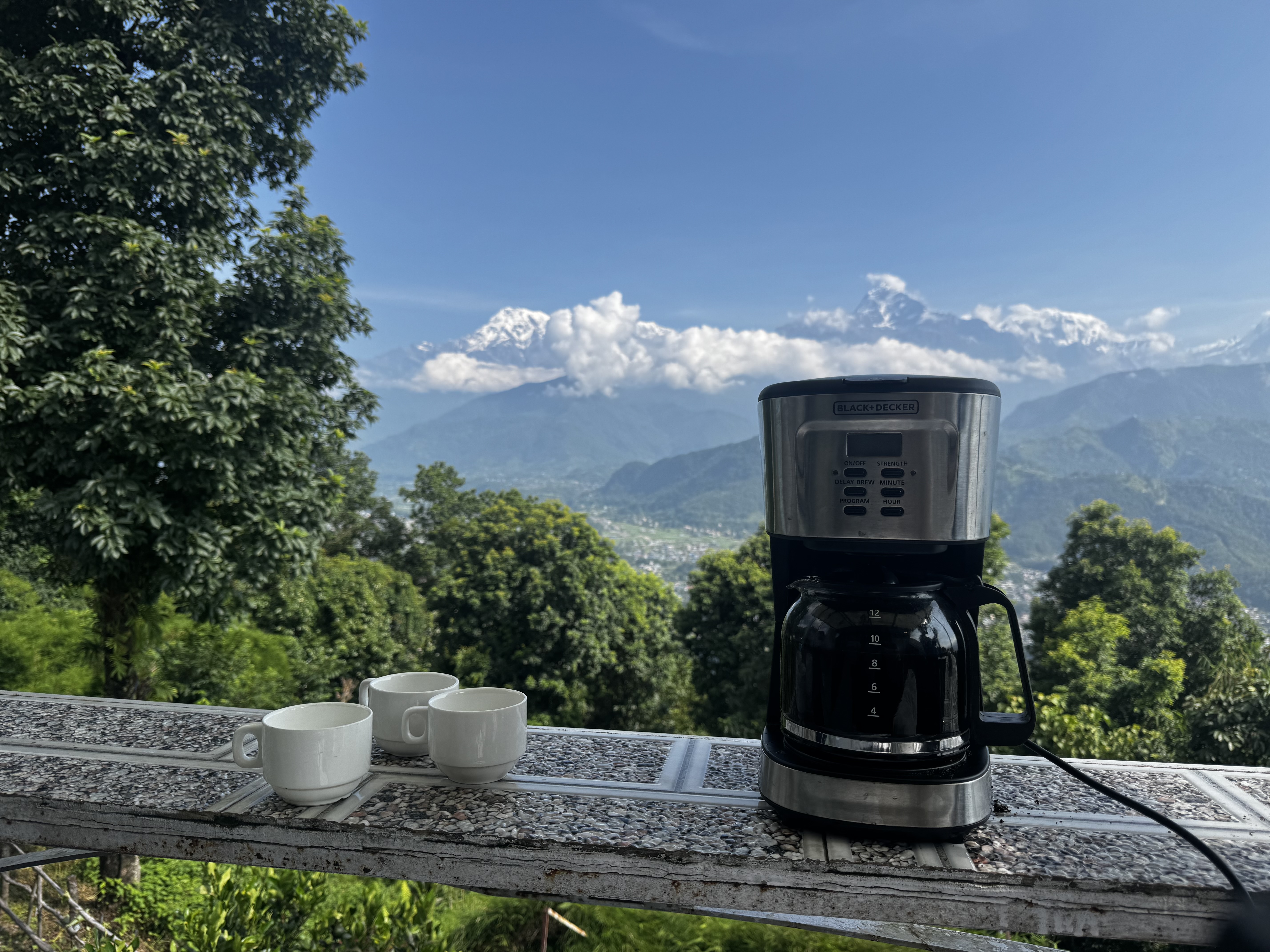 A coffee maker sits on a tiled outdoor table with three white cups beside it, overlooking a scenic view of mountains and clouds. The lush greenery in the foreground adds depth to the landscape, with the distant peaks partially covered in snow under a bright blue sky.