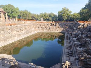 A serene view of a stepwell surrounded by intricately carved stone steps and walls. The water in the stepwell is still and reflects the clear blue sky and surrounding greenery. Some trees are visible in the background, and a pathway made of wooden railings runs along the edge.
