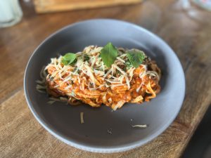 A close-up of a popular dish of Spaghetti Bolognese on a dark gray plate. The pasta is coated in a reddish-orange sauce and topped with shredded cheese and fresh green basil leaves. The plate sits on a wooden table
