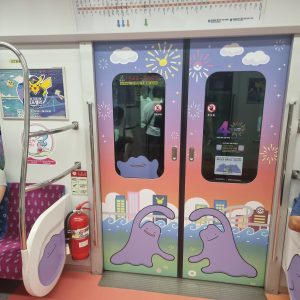 A colorful subway train door featuring playful cartoon-style designs of two purple, blob-like creatures with smiling faces. The door is set against a gradient background of soft orange and blue, decorated with clouds, stars, and fireworks. Nearby, a metal pole and seat can be seen, along with a fire extinguisher and a train map on the wall, indicating a lively and vibrant atmosphere inside the train.
