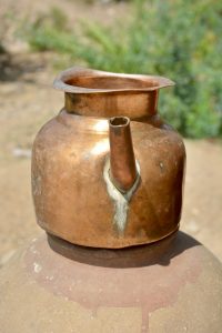 A traditional copper kettle with a weathered patina, placed under bright sunlight, showcases its rustic charm and timeless craftsmanship. Captured from Bhangarh Fort, Alwar, Rajasthan.