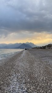A wide pebble beach at sunset with calm waves lapping the shore, backed by a dramatic mountain range under a cloudy sky. Palm trees and buildings line the right side of the coastline in the distance. Konyaaltı Beach, Antalya, Turkey