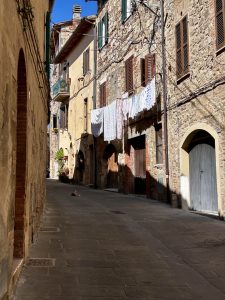 A quiet summer day in a small Italian town, with narrow cobblestone streets, rustic stone buildings, wooden shutters, and laundry hanging to dry under the bright blue sky.