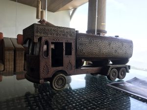 A decorative wooden model of a truck, intricately carved with floral patterns and designs. The truck has a rounded cab with an open front and four wheels. It features a cylindrical tank shaped body and is placed on a glass table, reflecting its surroundings. In the background, there are hints of wicker furniture and a view outside.