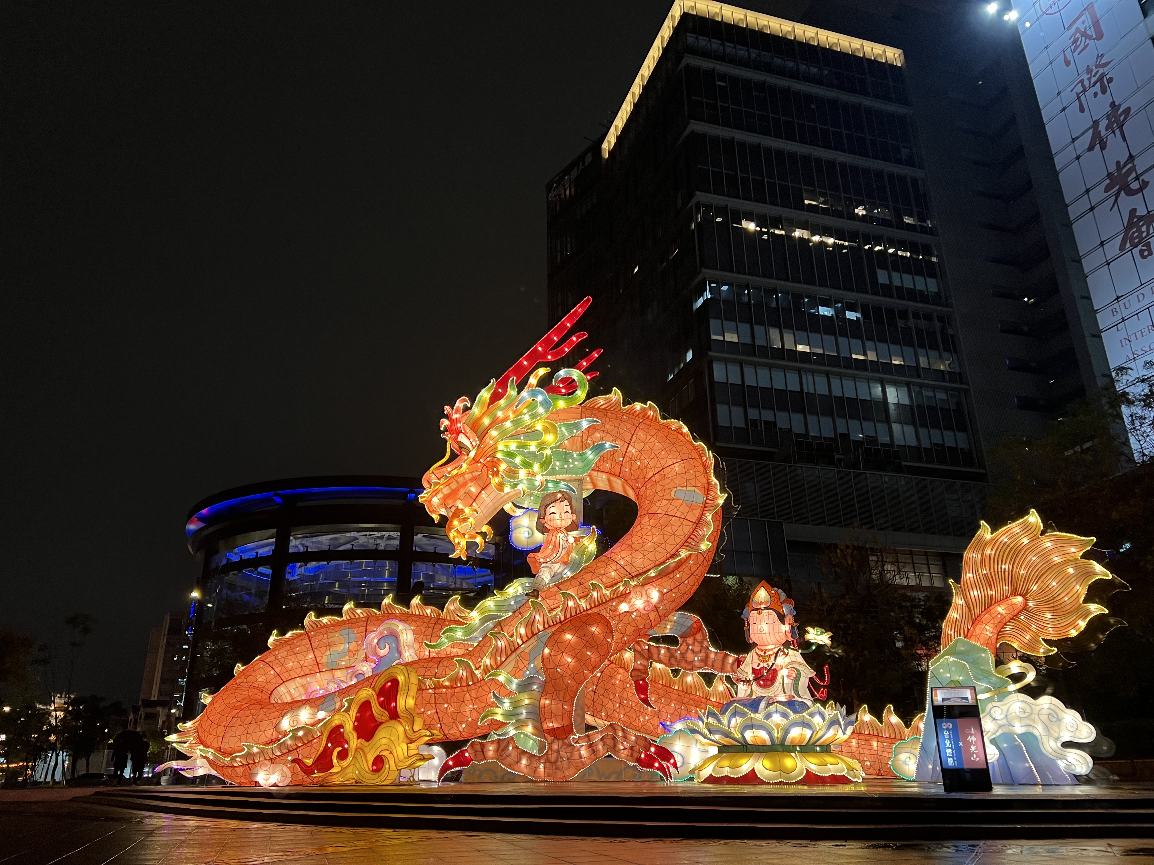 A large, illuminated dragon lantern with two figures riding on it, set up at night in an urban plaza with a modern building in the background from Taiwan