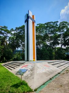 Shaheed Minar, a tall monument with white, orange, black, and green colors, stands proudly against a clear blue sky, surrounded by green trees.