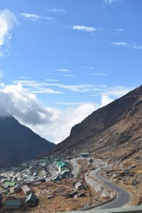 This photograph captures the scenic winding mountain road leading to Tsomgo (Changu) Lake, located in the Gangtok district of Sikkim, India. Nestled at an altitude of about 3,753 meters (12,313 ft), this route is known for its sharp bends, alpine landscape, and spectacular views of the Eastern Himalayas. The charming green-roofed houses of the near
