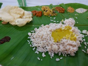 A top-down view of a traditional South Indian meal served on a banana leaf. The meal consists of white rice, sambar, various curries, and a crispy papadam.