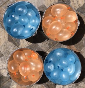 Four metal buckets filled with water balloons sit on a stone surface. Two buckets contain blue water balloons, while the other two have orange water balloons.