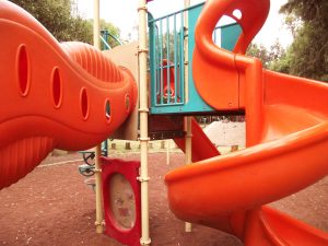 A close-up view of an orange playground slide and tunnel, featuring a textured surface and several circular openings. The slide connects to a multicolored play structure with a teal section above.