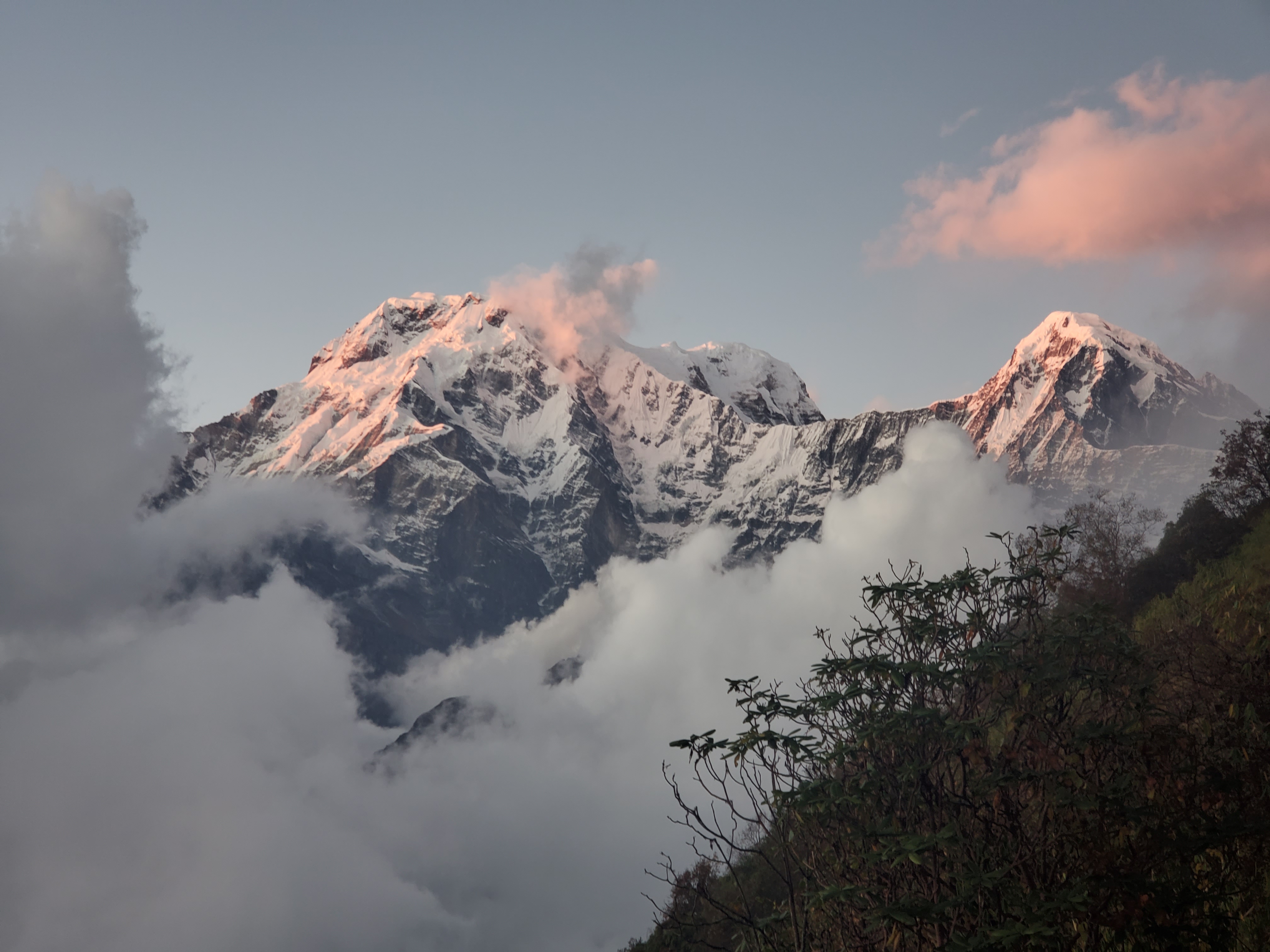 A mountain range with snow-covered peaks is partially obscured by clouds. The setting sun casts a soft, pink glow on the tops of the mountains and the surrounding clouds.