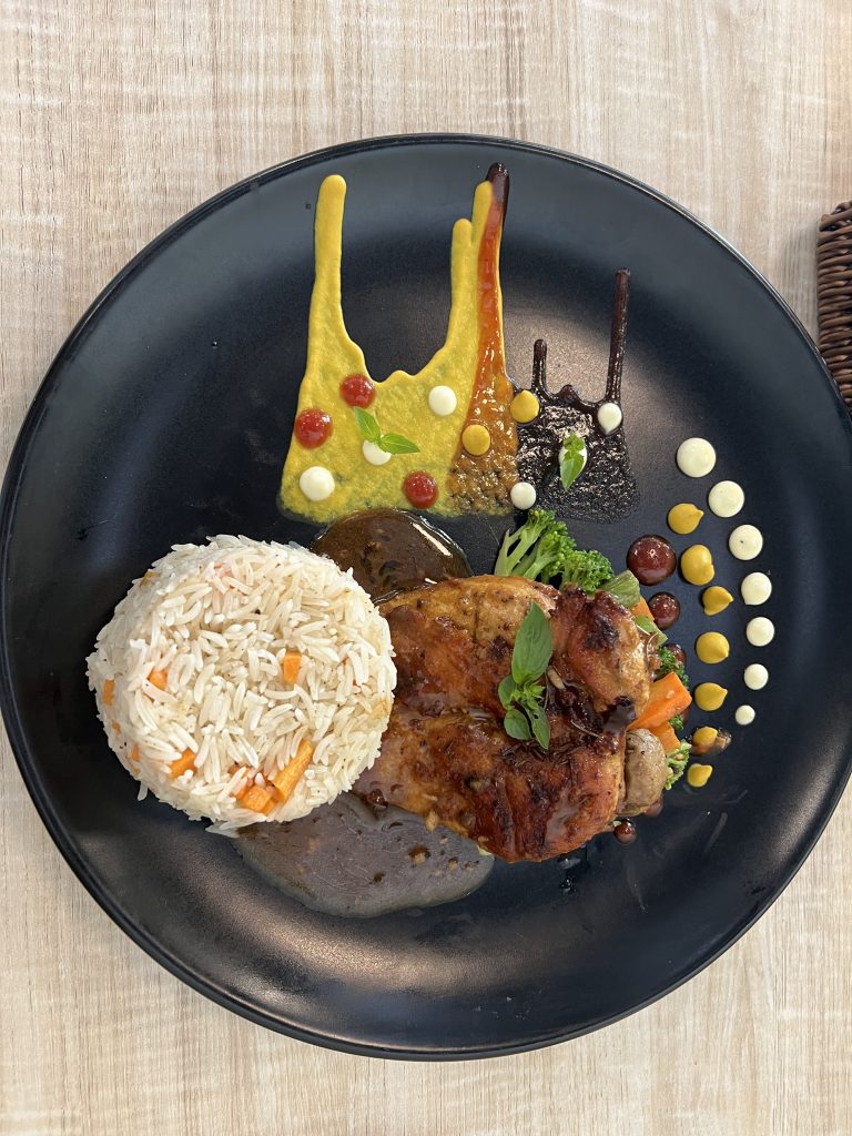 A gourmet dish with grilled chicken, rice, and vegetables, artistically plated with various sauces and dots of cream.