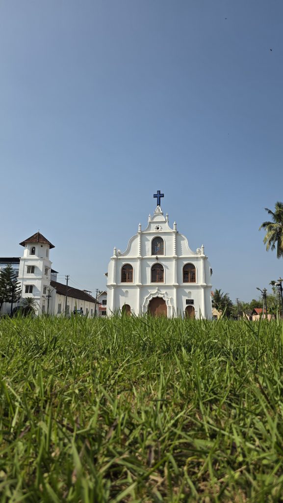 A white Catholic church with a prominent cross on its facade, standing amidst green grass under a clear blue sky, with a bell tower visible to the left.