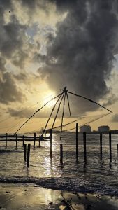 Chinese fishing nets silhouetted against a golden sunset over the backwaters of Fort Kochi, Kerala, with reflections shimmering on the water and a cloudy sky above.