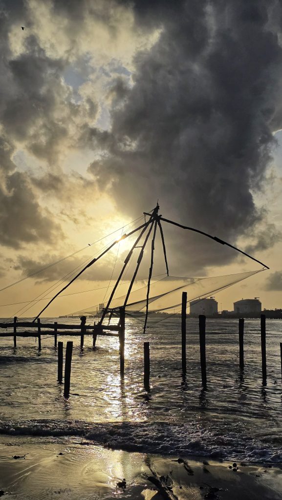 Chinese fishing nets silhouetted against a golden sunset over the backwaters of Fort Kochi, Kerala, with reflections shimmering on the water and a cloudy sky above.