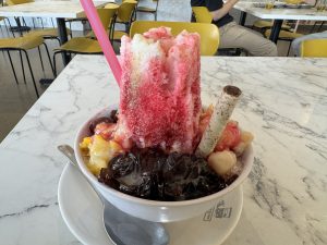 A big shaved ice in a white bowl with red syrup, fruit, jelly, a wafer stick, and a pink straw on top.