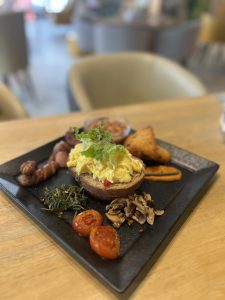 A black plate of a full breakfast with Brown bread, scrambled egg, sausages, bacon, baked beans, and other items.