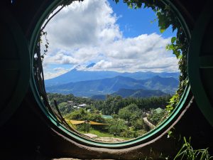 A circular window framed by greenery reveals rolling hills and mountains beneath a partly cloudy sky, blending nature and rustic charm.
