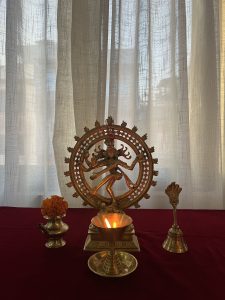 A statue of the god Nataraja on a red table. In front of it are a lit oil lamp, a small pot with an orange flower, and a brass bell. #SummerPhotoContest