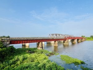 A red and grey truss bridge spans a wide river, supported by several concrete pillars. The water is calm, reflecting the blue sky and clouds above. Lush green vegetation lines the riverbank, with some water plants visible in the shallow areas near the shore.