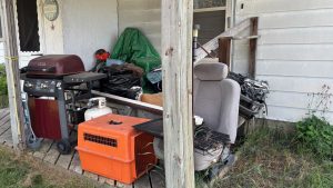 On a small porch, a grill, a chair from a car, a dog coop, and some other things.