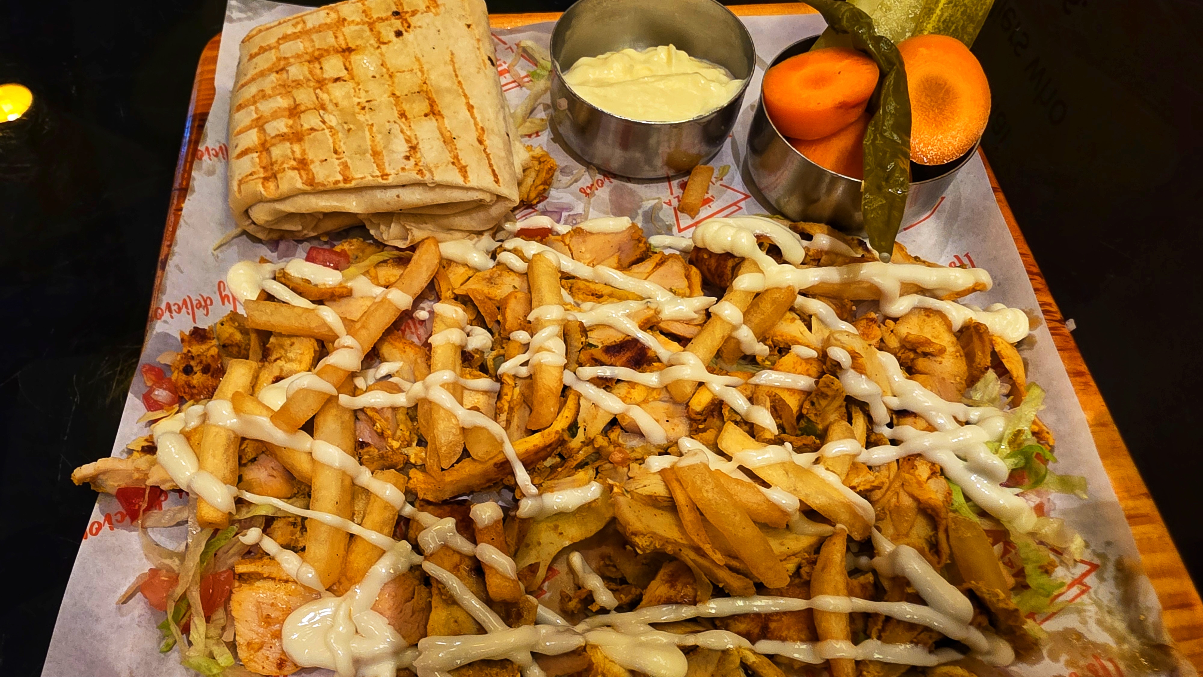 A top-down view of a rectangular platter filled with chicken shawarma, French fries, and two small white bowls containing dipping sauces, possibly garlic sauce and a chili sauce.