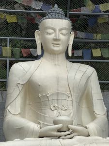 A full-body statue of a seated Buddha with a bowl in its lap, set against a wire fence with colorful prayer flags.