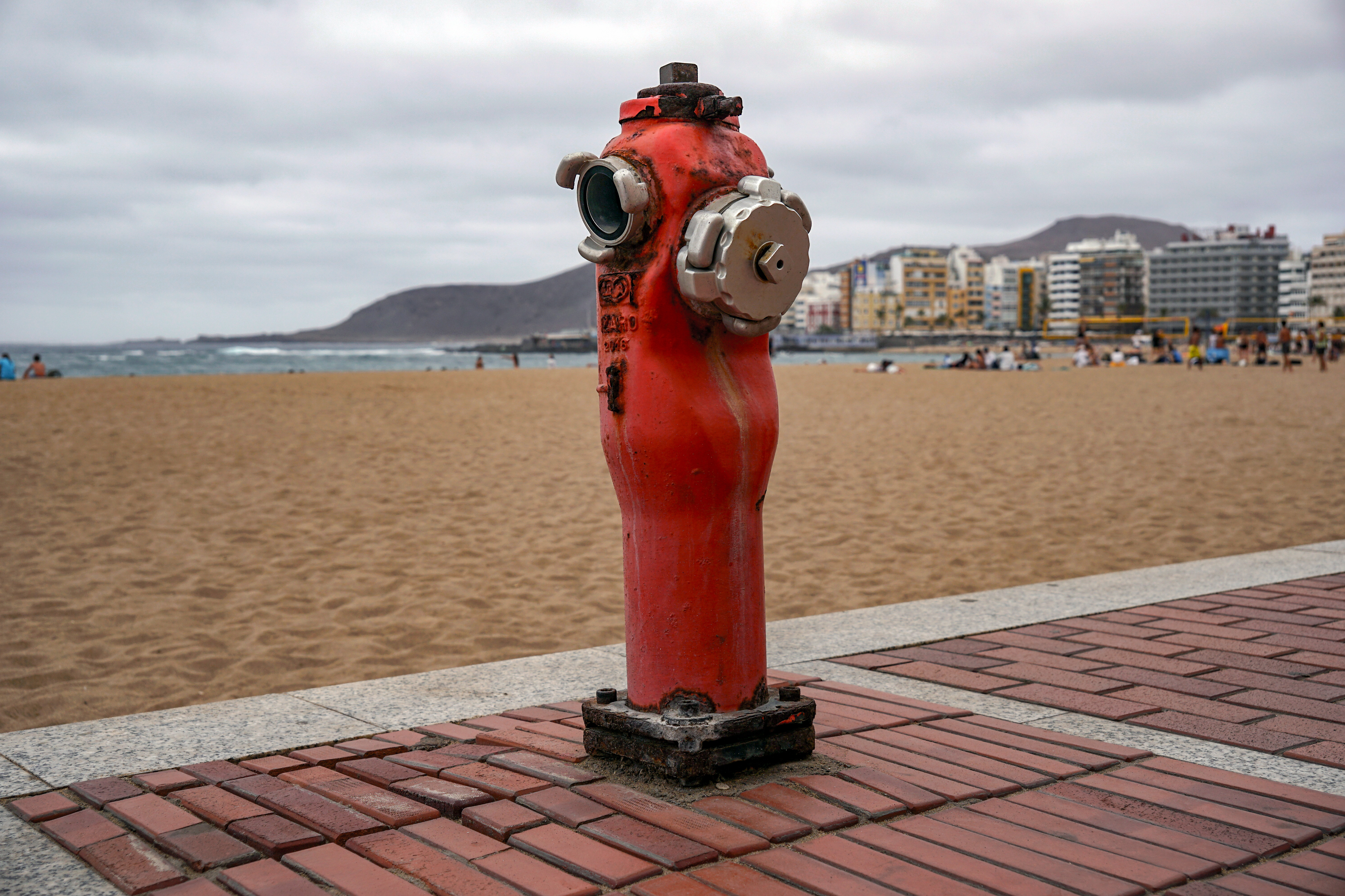 A bright red fire hydrant stands prominently on a brick walkway, overlooking a sandy beach. In the background, the ocean is visible with gentle waves, and a slightly cloudy sky looms above.