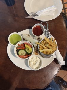 A plate of food on a wooden table. The plate contains a wrapped sandwich,a metal basket of French fries, a small bowl of salad, and three dipping sauces