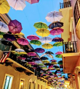 Colorful umbrellas at Calle de la Fortaleza in San Juan, Puerto Rico. A low angle view of orange, pink, purple and white umbrellas strung in an alley between beige buildings with cast-iron railed balconies.