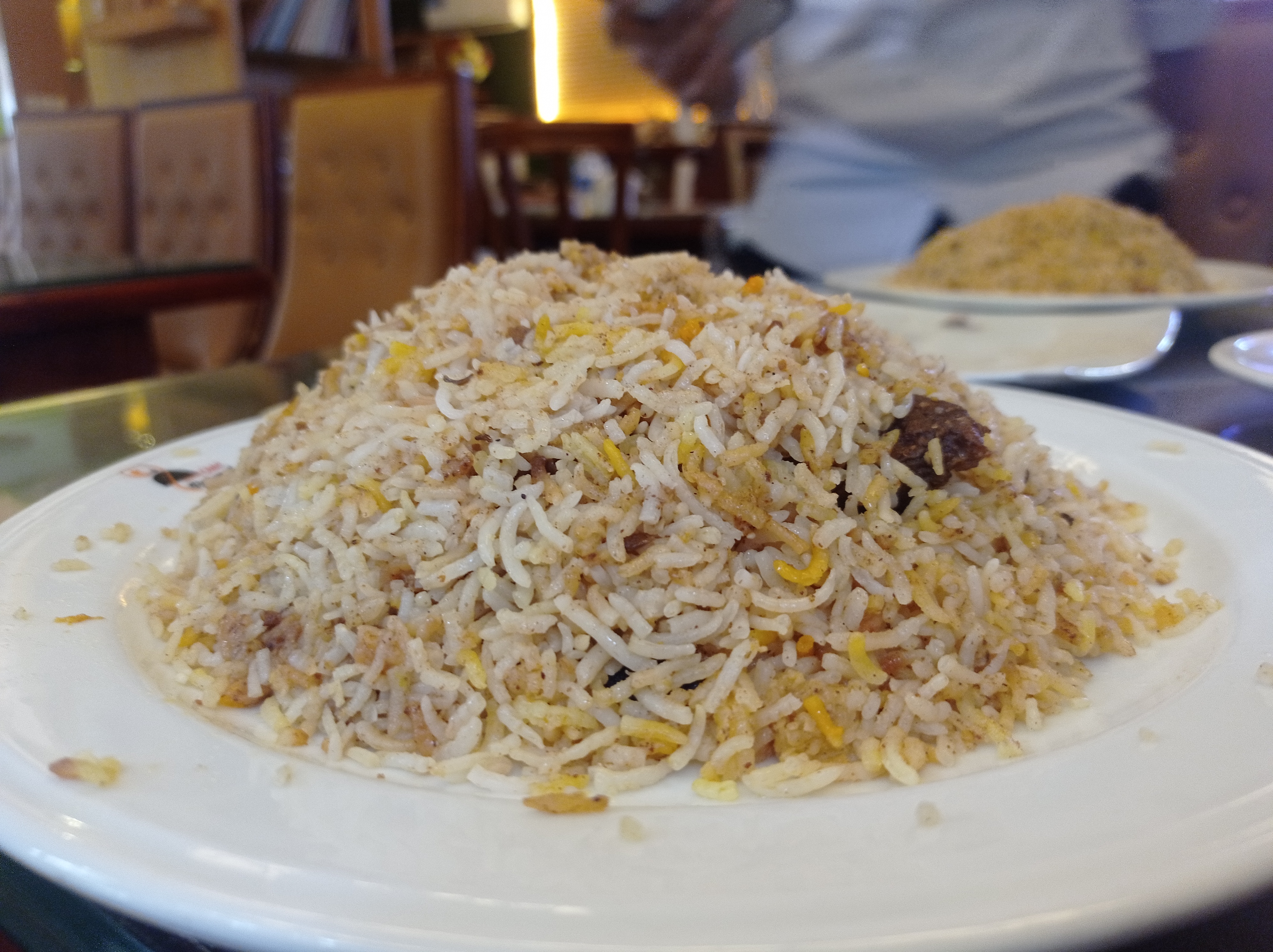 A close-up view of a large mound of biryani rice presented on a white plate. The rice is fluffy and golden, with specks of seasoning visible throughout.