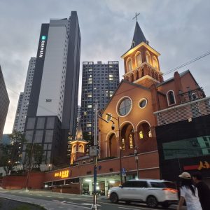 A lit-up brick church with arched windows and steeple stands on a street corner at dusk, surrounded by tall modern apartment buildings in Busan. Cars and pedestrians pass by in the foreground. Onchen Roman Catholic Church