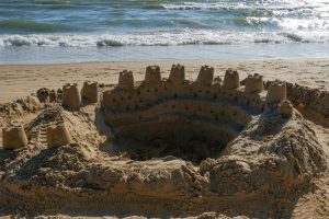 A circular sandcastle structure is built on a beach, featuring several towers with small windows and a moat-like depression in the center. The waves of the ocean gently lap at the shoreline in the background