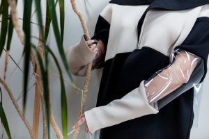 A fashion editorial. A close-up of a person wearing a modern black and beige outfit with oversized sleeves and transparent plastic arm coverings, standing next to a plant and lightly holding its branches.