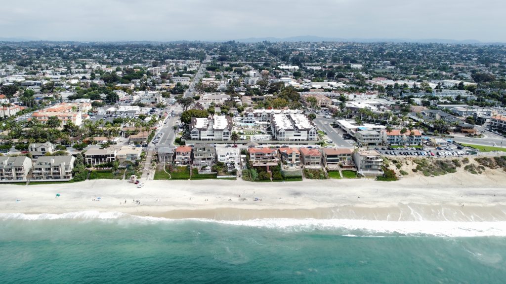 An aerial view of a coastal area featuring a sandy beach with gentle waves lapping at the shore. In the foreground, several beachgoers are seen relaxing on the sand, while beach umbrellas are set up. Behind the beach, there are rows of multi-story residential buildings and businesses. The area is lush with greenery, with palm trees interspersed among the structures. A road runs parallel to the beachfront, and the background shows a sprawling urban landscape with additional buildings and greenery under a partly cloudy sky.