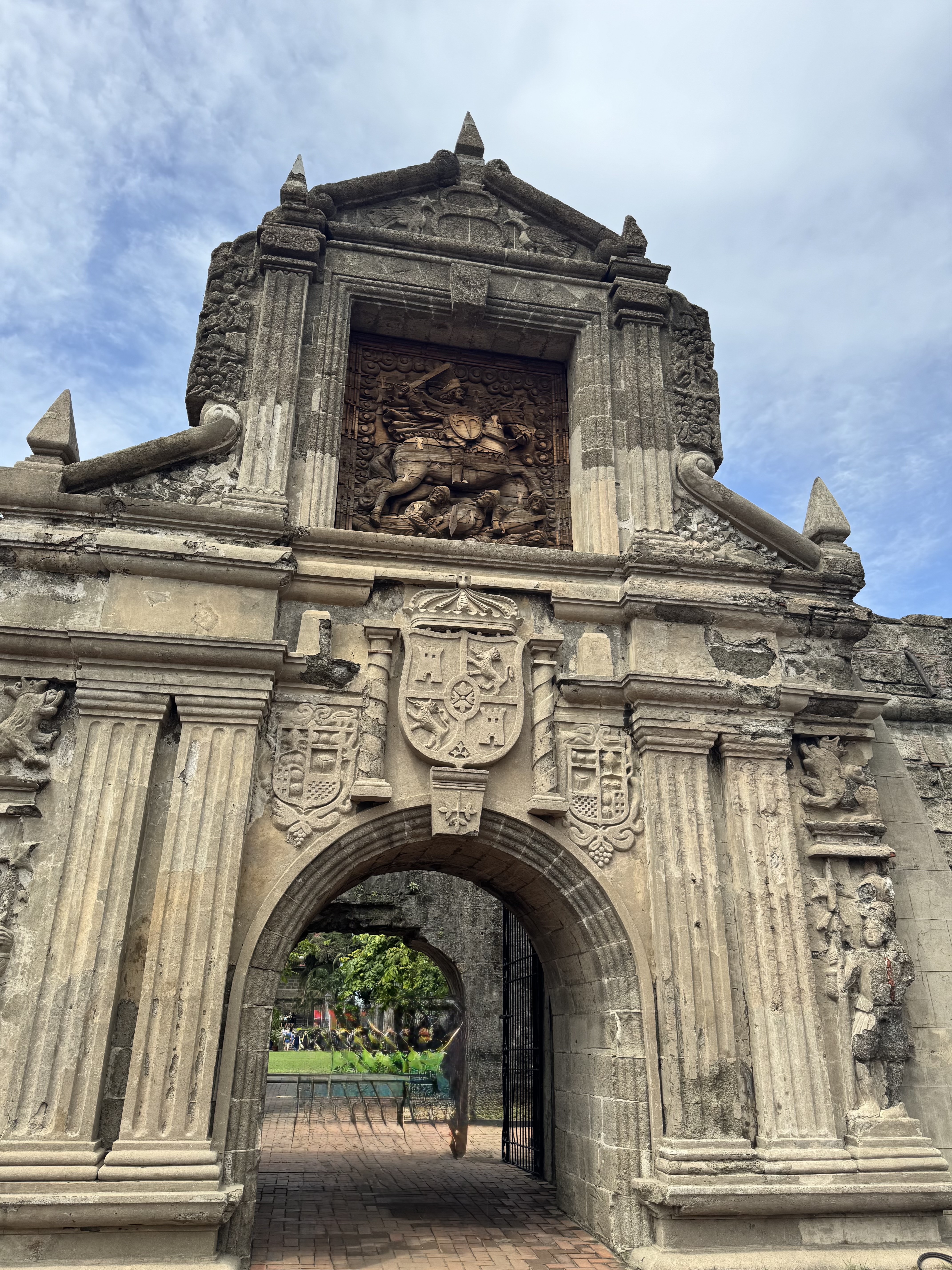 Fort Santiago’s stone archway with a bas-relief of a horseback rider fighting a dragon, leading to a tree-lined brick path under a cloudy sky.