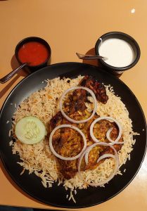 A single serving of chicken Mandhi is presented on a black plate with a side of cucumber, onion rings, and two dipping sauces, one red and one white. 