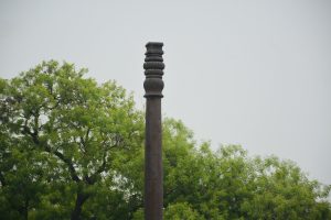 A detailed view of the top portion of the iconic Iron Pillar at the Qutub Complex, Delhi. Known for its rust-resistant composition, this ancient structure stands tall against a backdrop of fresh greenery, showcasing India’s rich metallurgical heritage. 
