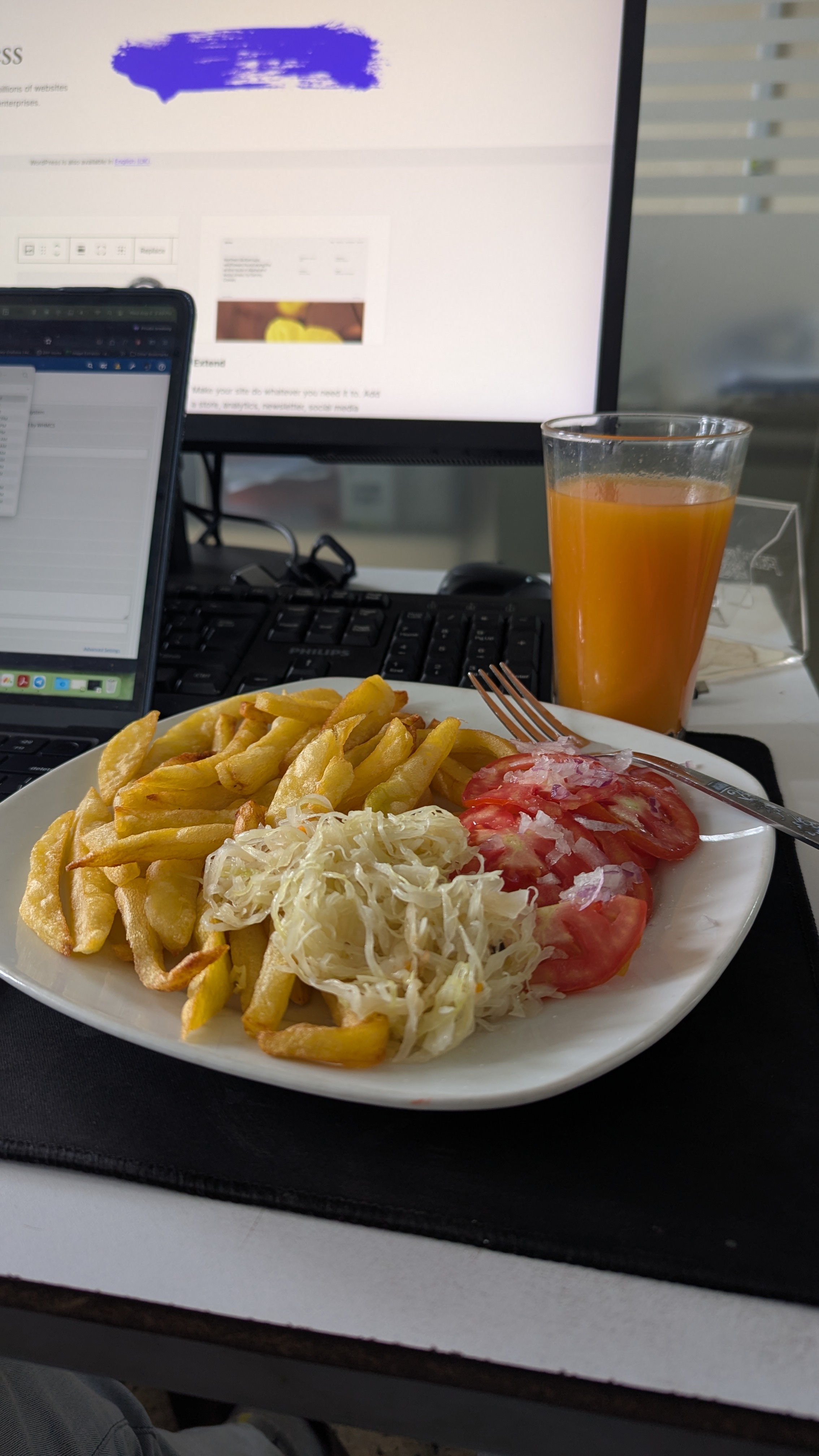 A plate of food featuring golden fried potatoes, a serving of shredded cabbage, slices of fresh tomatoes topped with diced onions, and a glass of orange juice. In the background, a computer monitor displaying a website is visible, along with a keyboard.