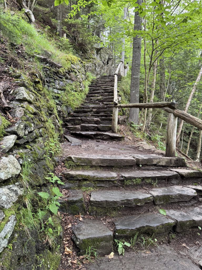 A rugged outdoor staircase with a wooden railing found on Glen Ellis Falls Trail in Gorham, New Hampshire along a mossy stone wall.