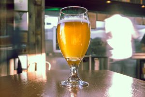 A tulip glass of hazy golden beer sits on a wooden table in a sunlit brewery taproom, with blurred figures and brewing equipment in the background. 