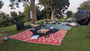 A nicely setup movie ares for group movies. Various patterned rugs are laid out, some in shades of red and teal, with black fabric and plush cushions. Each setup includes wooden pallets as tables, adorned with vases of flowers.