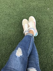 A low-angle shot of a person's legs crossed, wearing ripped jeans and sneakers with a pink and blue stripe, on a green artificial turf.