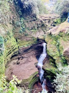 A narrow, rocky canyon filled with lush greenery shows a small waterfall cascading through the jagged cliffs. 