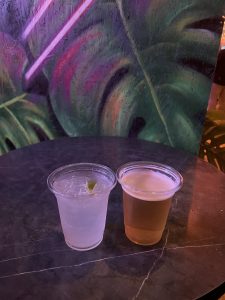 A clear plastic cup containing a light-colored drink with ice and a lime wedge sits next to a second clear plastic cup filled with a golden amber beverage. Both cups are placed on a dark table surface, with a colorful, vibrant mural featuring leaf patterns in the background and subtle neon lighting adding an atmospheric glow.