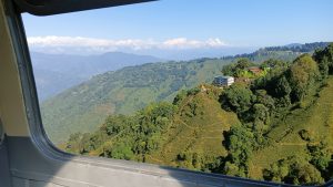 A view from the window of a plane shows lush green hillsides with terraced fields and scattered houses. In the background, there are distant mountains under a clear blue sky with some clouds.