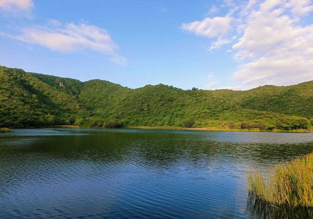 A serene landscape featuring a calm lake surrounded by lush green hills under a partly cloudy blue sky.