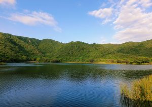 A serene landscape featuring a calm lake surrounded by lush green hills under a partly cloudy blue sky.
