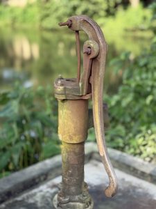 A close-up of an old-fashioned hand water pump, displaying rust and wear, set against a blurred background of lush green foliage and water. 
