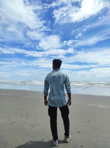 A man, seen from behind, stands on the sandy beach of Cox's Bazar, Bangladesh, looking out at the calm, blue-green ocean. He is wearing a patterned blue shirt and dark pants, with a wide expanse of beach in front of him and a dramatic sky filled with white clouds overhead.